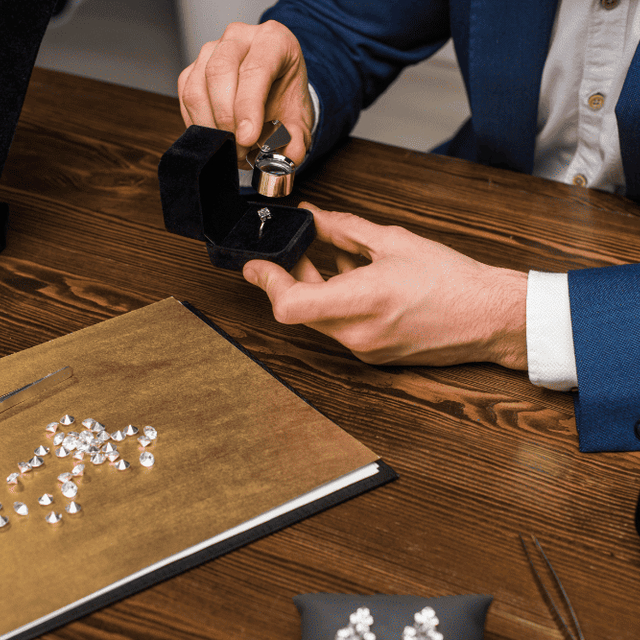 A jeweller presenting a ring in a box across a desk, with loose diamonds and tweezers to one side